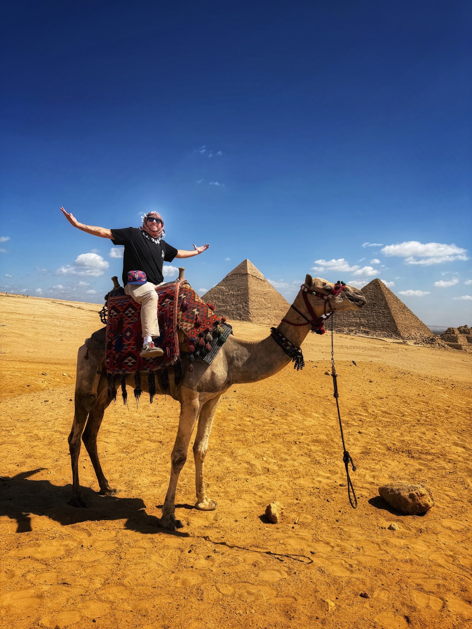 Ken with arms outstretched atop a camel before the pyramids of Giza.